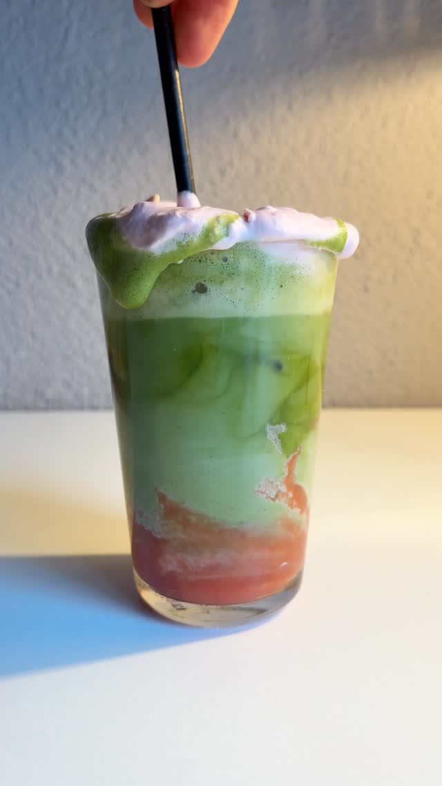 Glas mit Matcha Latte, Erdbeerschicht und rosa Milchschaum, ein Löffel rührt um